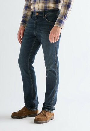 Mann trägt dunkelblaue Jeans, braune Wildleder-Mokassins und ein gelb-blau kariertes Langarmhemd, steht vor einem einfarbigen Hintergrund.