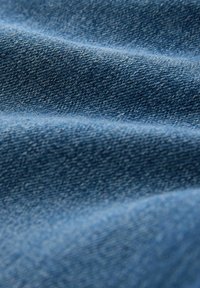 Denimstoff in einem mittleren Blauton, der eine strukturierte Webart mit subtilen Schattierungsvariationen zeigt. Weiches, fließendes Erscheinungsbild mit glatter Oberfläche.