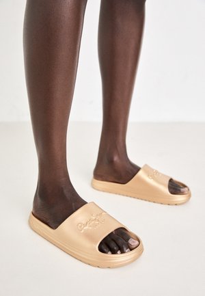 Sandalias de slide beige con diseño de punta abierta, textura suave y logo en relieve. Suela plana con un estilo minimalista.