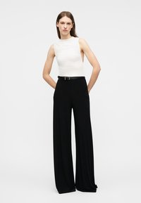 Γυναίκα που στέκεται με τα χέρια πίσω από την πλάτη, φορώντας αμάνικο λευκό τοπ και μαύρο παντελόνι με φαρδιά μπατζάκια και ψηλή μέση, με μαύρη ζώνη.