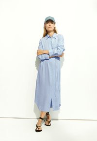 Robe chemise bleu clair à manches longues avec des rayures verticales blanches, fermeture à boutons sur le devant, et fentes latérales, associée à des sandales noires à lanières et une casquette.