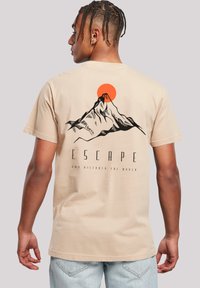 Young man with braided hair wearing beige T-shirt with mountain and red sun graphic and text "ESCAPE and discover the world" on back.