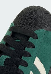 Groene suède sneaker met zwarte veters en een gestructureerde zwarte rubberen neuskap. Heeft witte zigzagstreep details aan de zijkant.
