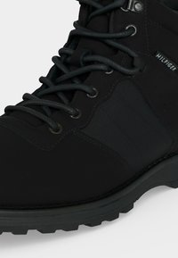 Gros plan sur une botte cheville noire avec un panneau latéral texturé, des œillets métalliques, des lacets ronds et une petite étiquette en tissu indiquant "Hilfiger".