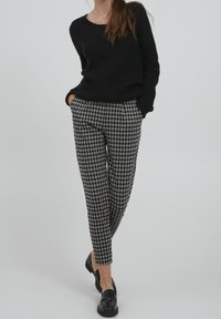 Zwarte gebreide trui met lange mouwen, gecombineerd met grijs- en zwartgeblokte broek. Zwarte instappers maken de outfit compleet.