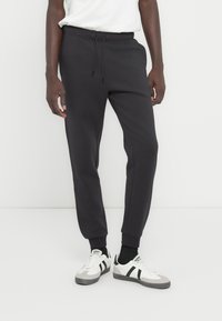 Σκουρόχρωμο γκρι παντελόνι φόρμας με στενό κατέβασμα στα πόδια, ελαστική μέση, ρυθμιζόμενο κορδόνι και ριμπ μανσέτες. Συνδυάζεται με άσπρα αθλητικά παπούτσια.