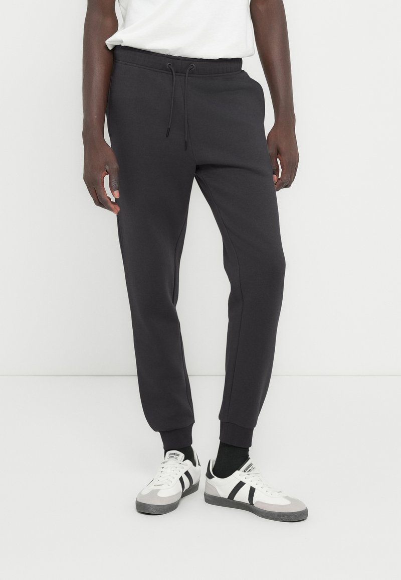 Σκουρόχρωμο γκρι παντελόνι φόρμας με στενό κατέβασμα στα πόδια, ελαστική μέση, ρυθμιζόμενο κορδόνι και ριμπ μανσέτες. Συνδυάζεται με άσπρα αθλητικά παπούτσια.