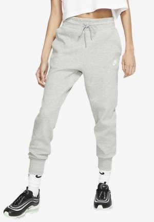 Femme portant un jogging Nike gris clair avec chevilles resserrées, des chaussettes blanches Nike, des baskets Nike noires et blanches, et un crop top blanc.