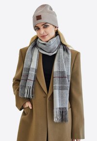 Écharpe à carreaux gris et blancs avec des franges, associée à un bonnet en tricot beige avec un patch en cuir marron, portée sur un haut noir et un manteau marron.