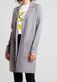 Abrigo largo de mezcla de lana gris con solapa, abertura frontal y dos bolsillos frontales. Lleva una camiseta con gráfico y pantalones negros.