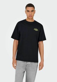Sort bomulds t-shirt med korte ærmer, prydet med gul tekst på brystet. Rund halsudskæring og afslappet pasform. Kombination med lysegrå jeans.