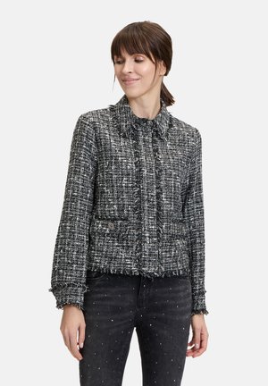 Schwarze und weiße Tweedjacke mit Fransen, Kragen, Fronttaschen und Knopfschluss. Kombiniert mit schwarzen, abgenutzten Jeans.