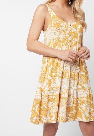 Robe jaune sans manches avec motif botanique blanc, devant boutonné et jupe à volants portée par une personne aux cheveux clairs.