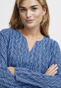 Blue patterned blouse featuring a wavy design in darker blue. V-neckline and long sleeves with a relaxed fit. Fabric appears soft.
