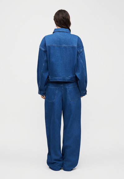 Άτομο με την πλάτη, φοράει τζιν μπουφάν και φαρδιά τζιν παντελόνια στον ίδιο μπλε χρωματισμό, σε λευκό φόντο.