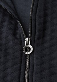 Närbild av en delvis dragkedjad svart quiltad tygjacka med en silverfärgad rund dragkedjehandtag.