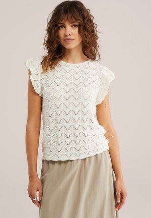 Kobieta o kręconych brązowych włosach, ubrana w biały koronkowy top bez rękawów z marszczonymi ramionami oraz beżową spódnicę na jednolitym tle.