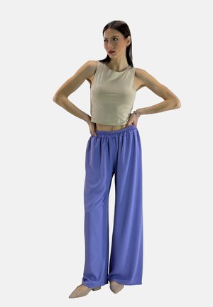 Beige cropped tanktop gecombineerd met losse, lichtpaarse broek met een elastische tailleband. Model staat met handen op de heupen.