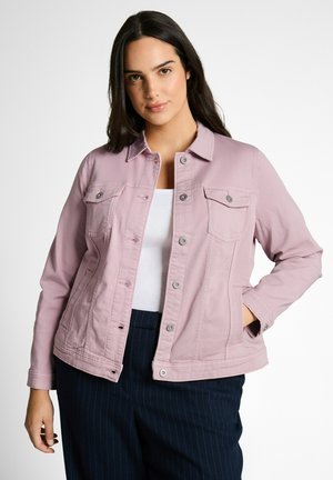 Junge Frau mit langen dunklen Haaren, die eine hellrosa Jacke mit Knöpfen über einem weißen Top und dunklen Nadelstreifenhosen trägt, posiert mit einer Hand in der Tasche.