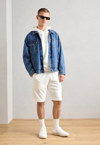 Jeansjakke i mellomblå, hvit hettegenser, teksturerte hvite shorts, hvite sneakers og solbriller. Funksjonelle lomme detaljer på jakken.