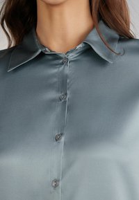 Camisa de satén en verde azulado con cuello, que presenta un cierre frontal con botones y una textura suave y brillante. Los botones son de un tono a juego.