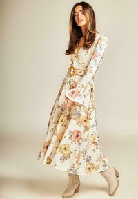 Vestido maxi floral de tela crema con mangas largas tipo campana y cintura ceñida, que presenta un diseño estampado de rosas y hojas; combinado con botas beige.