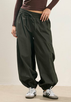 Person wearing dark green loose jogger pants with elastic cuffs and white sneakers with black stripes, standing against a plain wall.