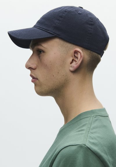 Junger Mann im grünen Hemd mit dunkelblauer Baseballkappe, seitlich vor einem hellen einfarbigen Hintergrund dargestellt.