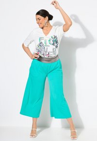 T-shirt bianco con scollo a V, stampa floreale e testo "FLOWER", abbinato a pantaloni palazzo teal e cintura argentata. Scarpe bianche con cinturini.