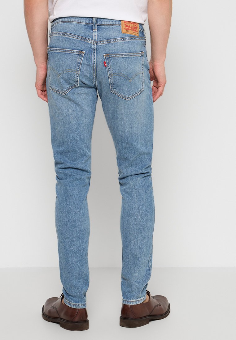 Homme portant un jean Levi's bleu clair et des chaussures habillées marron, se tenant de dos face à l'appareil photo sur un fond blanc.