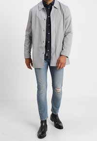 Ljusgrå vattentät jacka med krage, knappstängning och långa ärmar. Kombinerad med blå slitna jeans och svarta ankelstövlar.