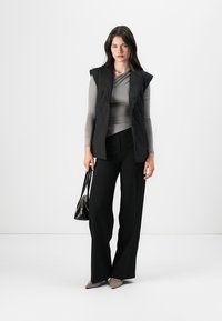 Gilet matelassé noir sur un haut gris à manches longues, associé à un pantalon large noir et des chaussures à talons pointus beiges, tenant un sac à main noir.