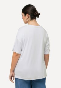 Mujer con el cabello oscuro recogido en un moño, usando pendientes de aro de plata, una camiseta blanca holgada y pantalones de mezclilla azul, vista desde atrás.