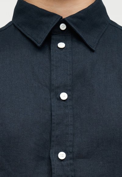 Κοντινό πλάνο μπροστινού μέρους ενός σκούρου μπλε πουκαμίσου με γιακά, με τέσσερα λευκά κουμπιά κλειδωμένα κατά μήκος της λωρίδας κουμπιών.