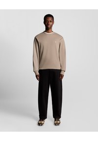 Langærmet beige sweatshirt med rund halsudskæring og subtilt logo, parret med sorte løse bukser og mønstrede brune og hvide sneakers.