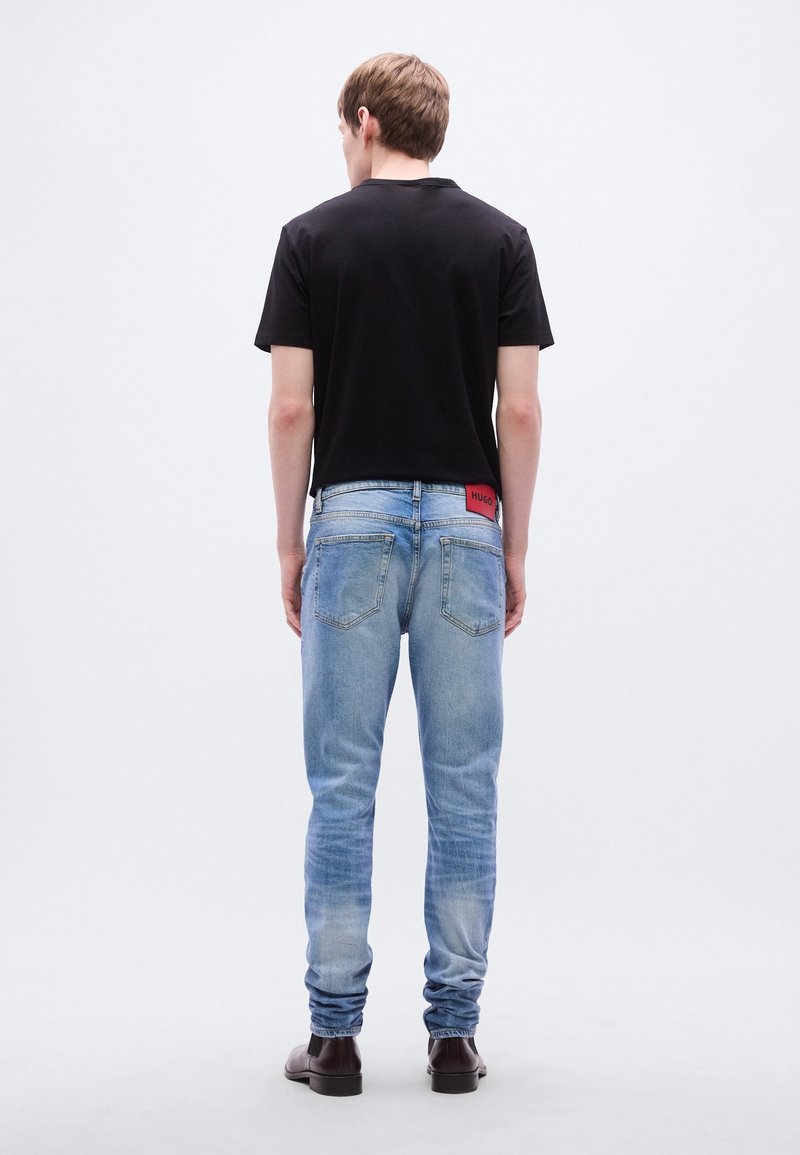 Jeune homme debout, de dos, portant un t-shirt noir, un jean bleu clair avec un patch rouge, et des chaussures marron foncé sur un fond blanc uni.