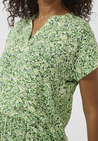 Groene blouse met bloemenprint en korte mouwen, voorzien van een V-hals, textuurstof en verschillende groene patronen op een witte achtergrond.
