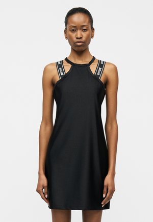 Žena v bezrukávových čiernych šatách s logovanými dvojitými ramienkami stojaca pred jednoduchým svetlým pozadím.