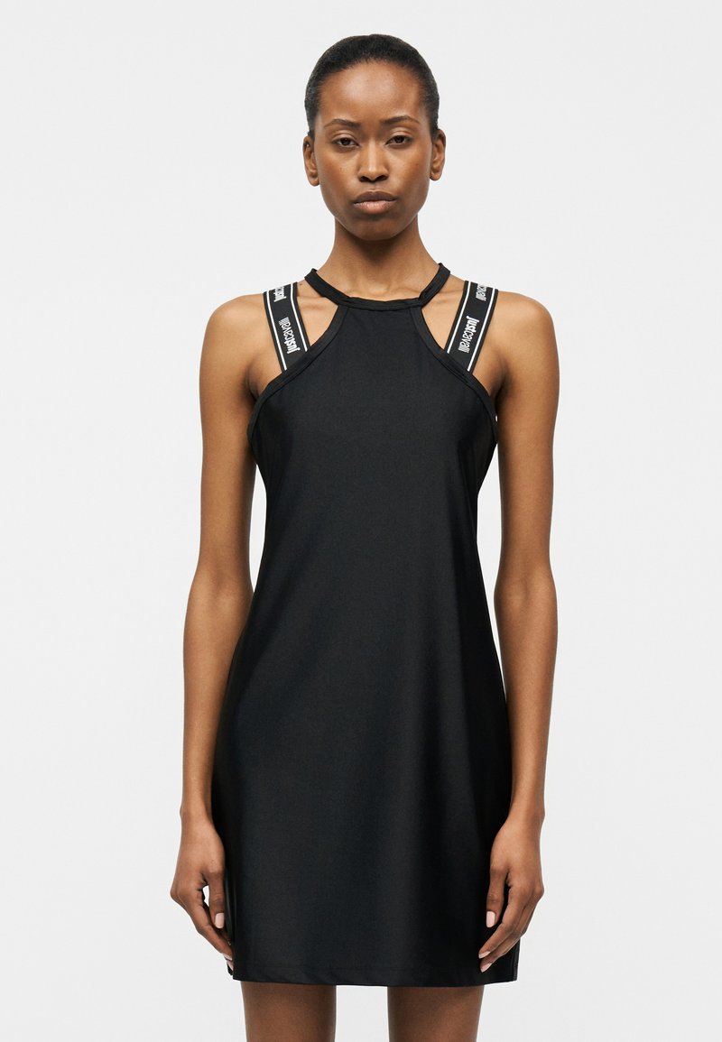 Γυναίκα που φοράει μαύρο αμάνικο φόρεμα με διπλές λουριά με λογότυπο στους ώμους, στέκεται μπροστά σε απλό ανοιχτό φόντο.