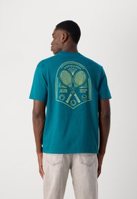 Teal bomulds T-shirt med en stor gul grafik af krydsede tennisracketer og tekst på ryggen. Korte ærmer og afslappet pasform.