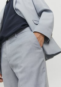 Lichtblauwe colbert en broek van een gestructureerde stof; aansluitend design; donkerblauw overhemd eronder; hand in de zak.