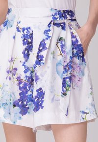 Shorts blancs à motifs floraux bleus et violets, avec une ceinture à nœud et des poches latérales pour une fonctionnalité supplémentaire.