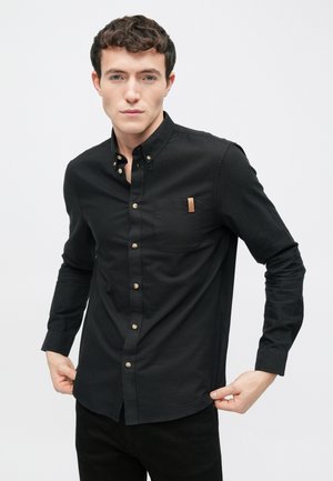 Chemise noire à boutons avec une poche poitrine ornée d'une étiquette beige, col standard, manches longues et tissu lisse et texturé.