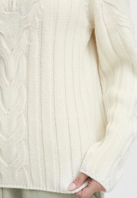 Pull en tricot blanc avec un motif en rib et une coupe décontractée. Le design présente des motifs torsadés et une finition douce.
