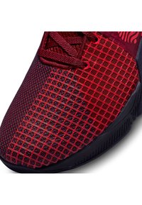 Gros plan d'une chaussure de sport en mesh rouge et noir avec un motif quadrillé en surimpression et des lacets bordeaux sur la partie supérieure.