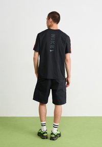 Černé tričko Nike s červenými akcenty, s vertikálním logem na zadní straně. Nosí se s černými cargo šortkami a botami s zelenými detaily.