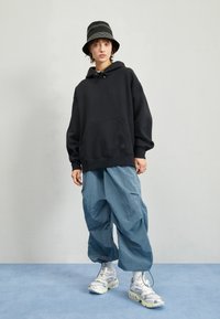 Sudadera oversized negra con bolsillo frontal, combinada con pantalones anchos azul claro y zapatillas blancas. Los accesorios incluyen un sombrero tipo bucket de rayas negras.
