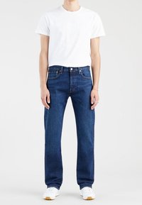Personne portant un t-shirt blanc uni, un jean bleu droit et des baskets blanches, debout devant un fond gris clair.