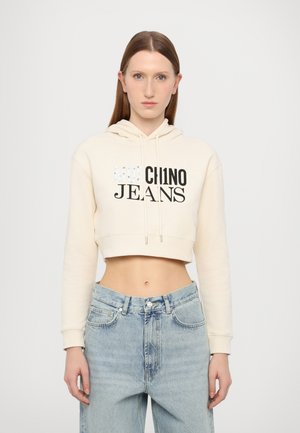 Cream cropped hoodie with front black text "Mochino Jeans," featuring a drawstring hood and soft material. Paired with high-waisted light denim jeans.