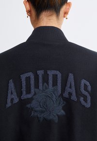 Zwarte jas met geborduurde "ADIDAS" tekst en een rozenontwerp op de achterkant; ribbelkraag en de stof heeft een gladde textuur.
